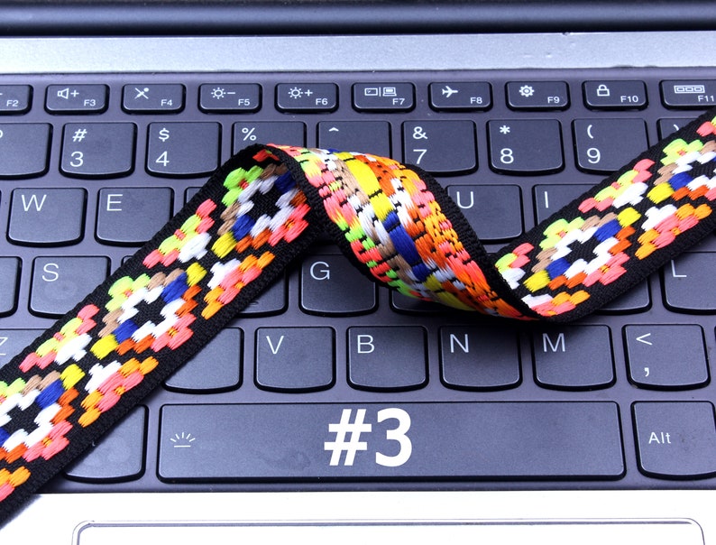 Op de afbeelding: Een kleurrijk geweven band met zwarte, witte, rode, oranje, gele, blauwe en groene accenten. Het band ligt op een zwart laptoptoetsenbord met het nummer "#3" zichtbaar op een van de toetsen.