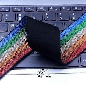 Pu&ograve; includere: Finitura elastica multicolore con finitura glitter. Il bordo ha strisce blu, verdi, argentate, arancioni e rosse. Il bordo &egrave; piegato a met&agrave; e posizionato su una tastiera di laptop nera. Il testo #1 &egrave; visibile sul laptop.