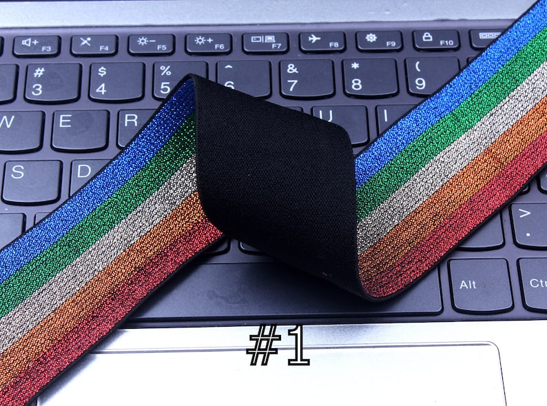 Pu&ograve; includere: Finitura elastica multicolore con finitura glitter. Il bordo ha strisce blu, verdi, argentate, arancioni e rosse. Il bordo &egrave; piegato a met&agrave; e posizionato su una tastiera di laptop nera. Il testo #1 &egrave; visibile sul laptop.