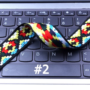 Op de afbeelding: Een kleurrijk geweven lint met zwarte, rode, gele, witte en blauwe strepen ligt over een laptoptoetsenbord. Het nummer "#2" is zichtbaar op het toetsenbord.