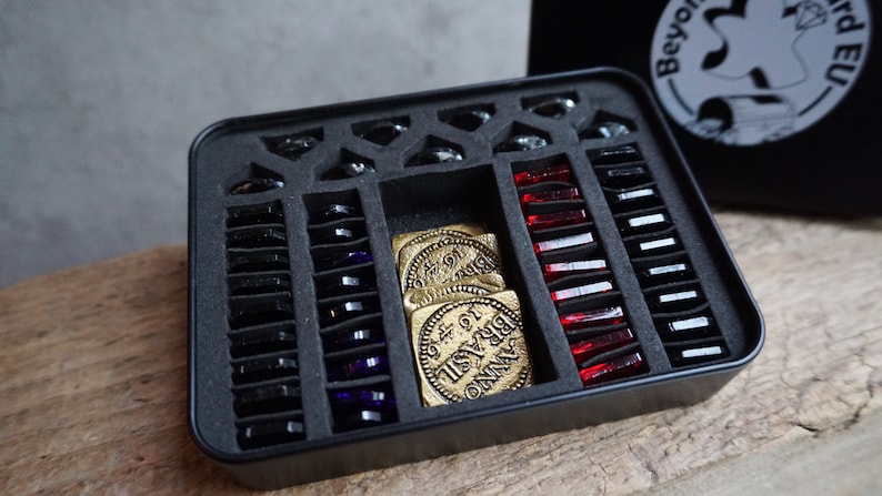 May include: A black metal box with a foam insert for storing game pieces. The box contains black, purple, and red glass pieces, as well as gold coins with the words "Anno Domini" and "BRYSAIL" on them.