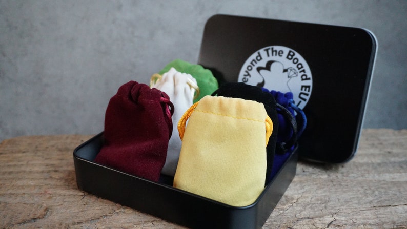May include: A black metal box with a white logo that says "Beyond The Board EU" and contains six small velvet drawstring bags in different colors: red, white, yellow, green, black, and blue.
