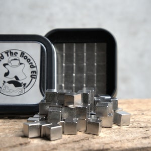 Puede incluir: Una lata de metal plateada con una tapa negra y el texto "Beyond The Board EU" en la tapa. La lata est&aacute; abierta y contiene un mont&oacute;n de peque&ntilde;os cubos de metal plateado.