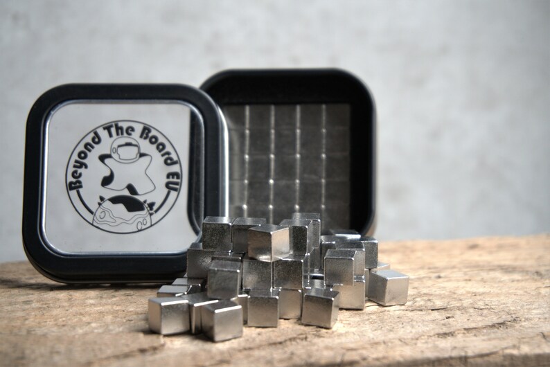 Puede incluir: Una lata de metal plateada con una tapa negra y el texto "Beyond The Board EU" en la tapa. La lata est&aacute; abierta y contiene un mont&oacute;n de peque&ntilde;os cubos de metal plateado.