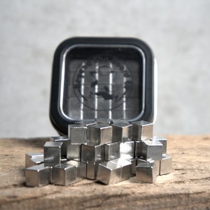 Puede incluir: Primer plano de un mont&oacute;n de peque&ntilde;os imanes en forma de cubo de color plateado. Los imanes est&aacute;n dispuestos en un mont&oacute;n suelto sobre una superficie de madera. Una lata negra con las palabras "Beyond The Board" es visible en el fondo.