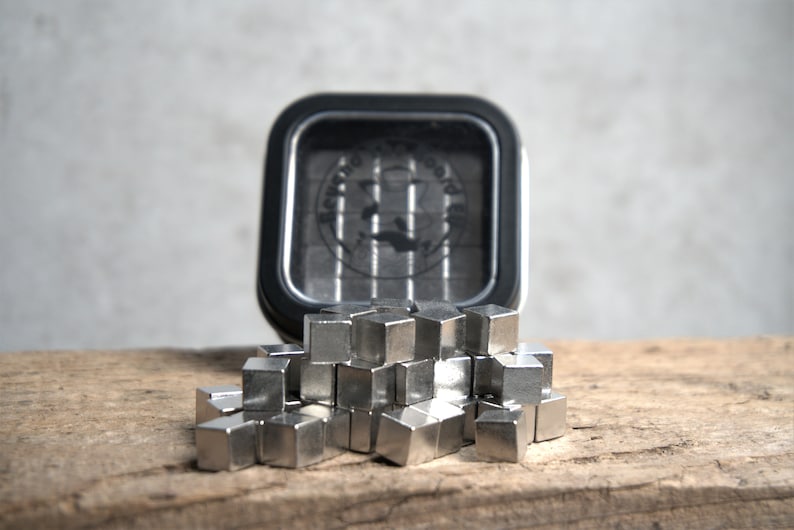 Puede incluir: Primer plano de un mont&oacute;n de peque&ntilde;os imanes en forma de cubo de color plateado. Los imanes est&aacute;n dispuestos en un mont&oacute;n suelto sobre una superficie de madera. Una lata negra con las palabras "Beyond The Board" es visible en el fondo.