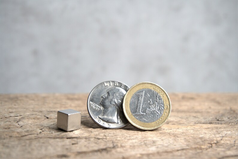 Puede incluir: Un im&aacute;n c&uacute;bico plateado junto a una moneda de cuarto de d&oacute;lar estadounidense plateada y una moneda de euro plateada con un anillo exterior dorado. Las monedas est&aacute;n sobre una superficie de madera.