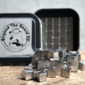 Puede incluir: Un conjunto de cubos magn&eacute;ticos plateados en un contenedor negro con el logotipo "Beyond The Board EU".