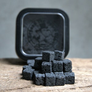 Puede incluir: Un contenedor negro con un logotipo blanco y negro, junto a una pila de pequeños cubos de carbón vegetal negros.