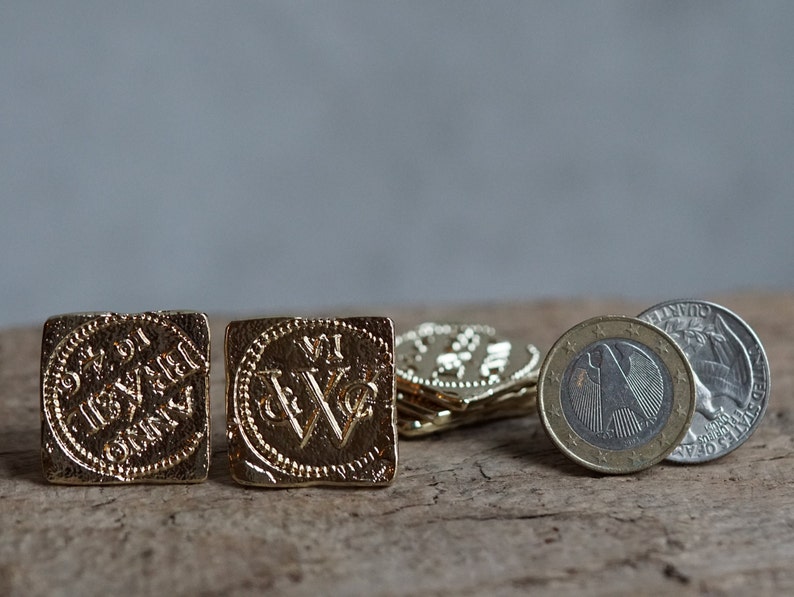 May include: Four gold-colored coins with intricate designs and lettering, including the words "S.R. DI" and "VI", are arranged on a wooden surface. A silver coin with a bird design is also visible.