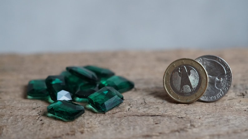 May include: A pile of dark green, faceted, rectangular glass gems on a wooden surface. Next to the gems are two coins, one silver and one gold.