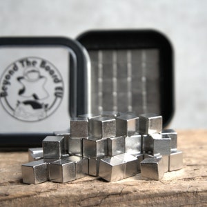 Puede incluir: Un contenedor de metal plateado con una tapa negra y el texto "Beyond The Board" en la tapa. El contenedor est&aacute; abierto y un mont&oacute;n de peque&ntilde;os cubos de metal plateado se est&aacute;n derramando.