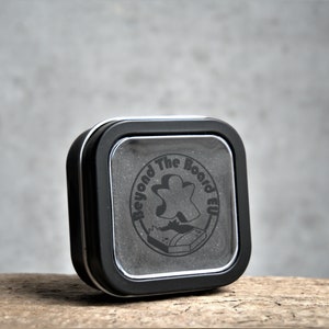 Puede incluir: Una lata cuadrada negra con una ventana de pl&aacute;stico transparente en la parte delantera. La lata tiene un logotipo en blanco y negro que dice "Beyond The Board EU" con un icono de pieza de juego.