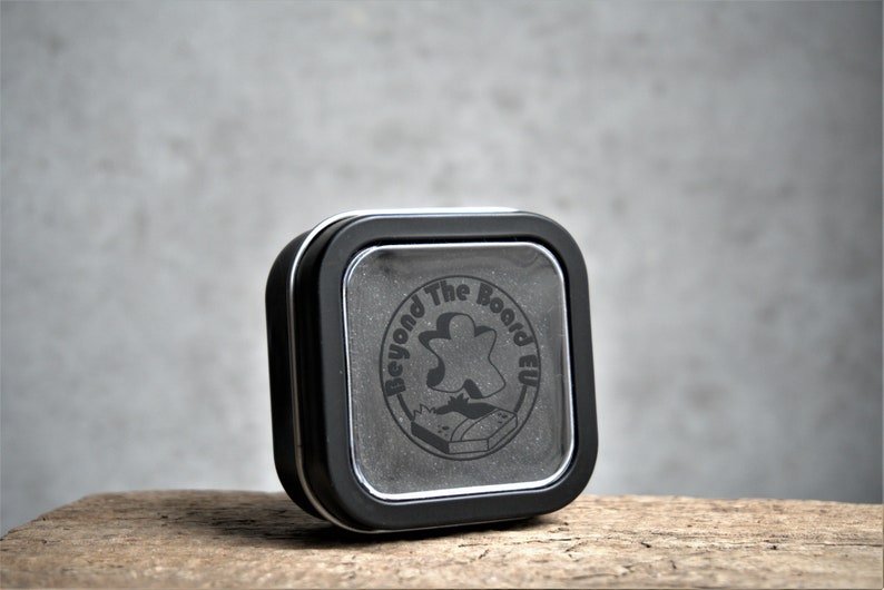 Puede incluir: Una lata cuadrada negra con una ventana de pl&aacute;stico transparente en la parte delantera. La lata tiene un logotipo en blanco y negro que dice "Beyond The Board EU" con un icono de pieza de juego.