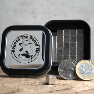 Puede incluir: Una peque&ntilde;a caja de hojalata con tapa negra y fondo de pl&aacute;stico transparente. La caja contiene un juego de peque&ntilde;os cuadrados magn&eacute;ticos plateados. La caja tiene un logotipo en la tapa que dice "Beyond The Board EU". Tambi&eacute;n hay dos monedas, una moneda de un cuarto de d&oacute;lar estadounidense y una moneda de euro, junto a la caja.