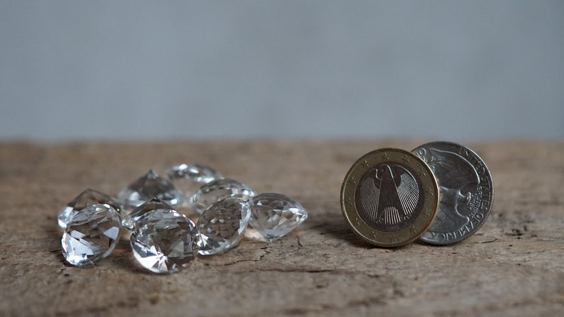 May include: A group of clear crystal beads, some with a faceted design, are scattered on a wooden surface.  Next to the beads are two coins, one a silver US quarter and the other a gold and silver Euro coin.