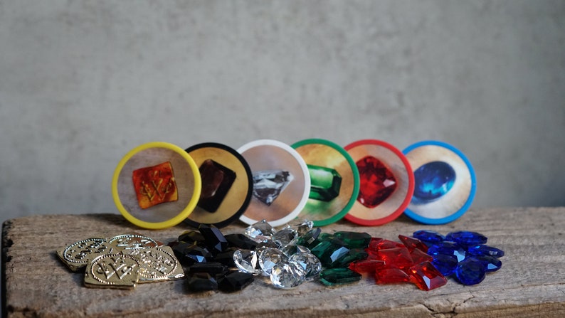 May include: A set of six colorful game tokens with different gemstones, including a red ruby, a blue sapphire, a green emerald, a diamond, a black onyx, and an orange amber. The tokens are surrounded by loose gemstones of the same colors.