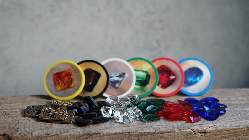 May include: A set of six colorful game tokens with different colored gems, along with piles of matching colored gems. The tokens are round and have a yellow, black, white, green, red, and blue rim.