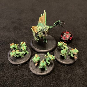 Puede incluir: Un conjunto de figuras de fantasía en miniatura, que incluye una gran criatura verde y naranja con aletas y un conjunto de criaturas verdes más pequeñas. Las figuras están sobre bases negras y hay un dado de 20 caras negro y rojo en el fondo.