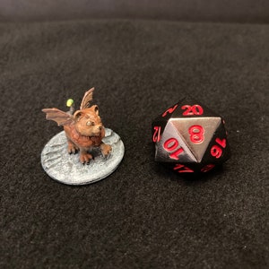Puede incluir: Un dado negro de 20 caras con números rojos, mostrando el número 16. Una pequeña miniatura de una criatura alada marrón sobre una base gris.