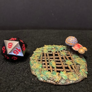 May include: A black 20-sided die with red numbers sits next to a miniature, hand-painted, circular, green and brown forest scene with a metal grate and a purple and orange mushroom.