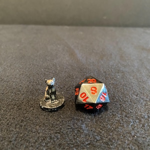 Puede incluir: Una pequeña figura de gato negro está junto a un dado de veinte caras. El dado es plateado con números rojos. La figura del gato está sobre una pequeña base redonda.