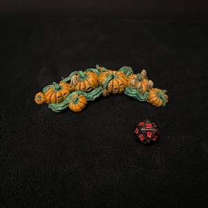 Puede incluir: Un grupo de dados en forma de calabaza naranja y verde con ojos negros, dispuestos en una pila sobre una superficie negra. Un dado negro de 20 caras con números rojos está en primer plano.