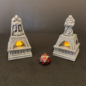 Peut inclure: Deux statues grises en 3D imprimées de personnages en capes, chacune debout sur une base avec une bougie allumée à l'intérieur. Un dé noir à 20 faces se trouve devant les statues.