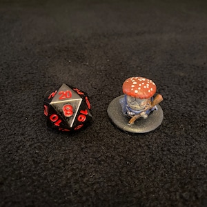 Puede incluir: Un dado negro de 20 caras con números rojos y una figura en miniatura de un personaje hongo con un sombrero de hongo rojo y blanco, ropa azul y un bastón.