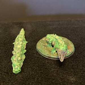 May include: Two green alligator figurines. One is a detailed, elongated alligator. The other is a miniature alligator emerging from a circular, green base. The alligators have textured skin and open mouths.