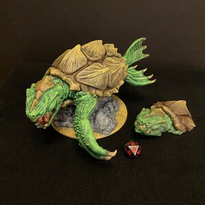 Pode incluir: Miniatura de fantasia detalhada, pintada à mão, de uma criatura semelhante a uma tartaruga com corpo verde e carapaça marrom. A criatura está em uma pequena base, com uma versão menor por perto. Um dado de vinte lados vermelho e preto também está presente.