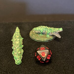May include: Three miniature crocodile figurines and a twenty-sided die. One crocodile is translucent green, another is green with an open mouth, and the die is black with red numbers. The items are arranged on a dark surface.