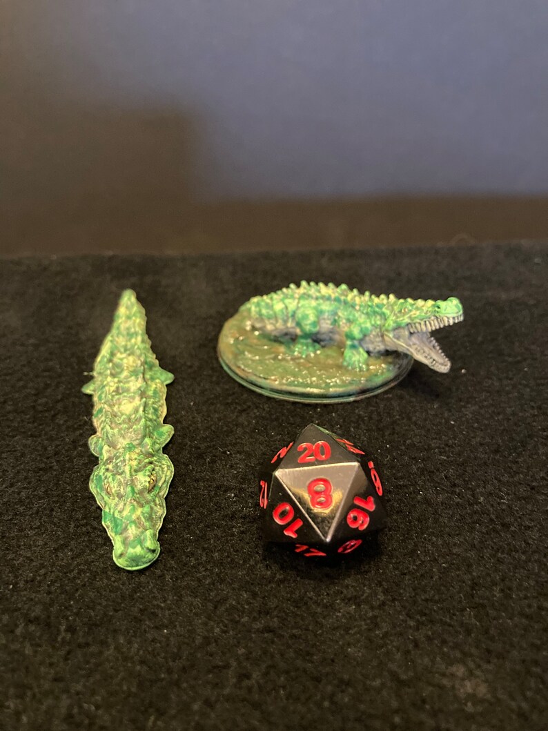 May include: Three miniature crocodile figurines and a twenty-sided die. One crocodile is translucent green, another is green with an open mouth, and the die is black with red numbers. The items are arranged on a dark surface.