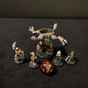 May include: A set of five miniature figures, including a large, metallic, barrel-shaped figure with four arms, and four smaller figures, all standing on black bases. A red and silver 20-sided die is in the centre of the image.