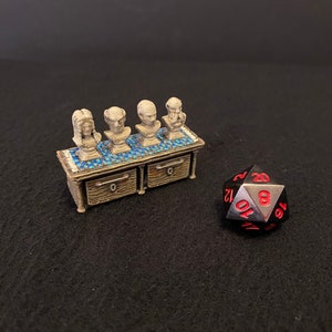 Könnte beinhalten: Ein Miniatur-Holzkabinett mit vier kleinen Büsten darauf. Das Kabinett hat zwei Schubladen und ist dunkelbraun lackiert. Die Büsten sind aus hellem grauen Material gefertigt und in einer Reihe angeordnet. Ein schwarzer 20-seitiger Würfel mit roten Zahlen befindet sich im Vordergrund.