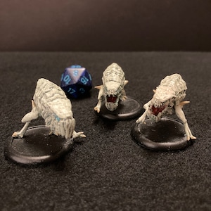 May include: Three white, skeletal, miniature figures with red mouths and blue eyes, standing on black bases. A blue and black 20-sided die is in the background.