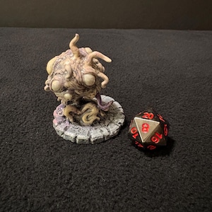 May include: A detailed, beige-colored fantasy creature figurine with multiple eyes and tentacles, sitting on a stone-like base. A twenty-sided die with red numbers rests beside it, suggesting a tabletop game theme.