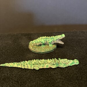 May include: Two miniature crocodile figurines. One is standing on a circular base, mouth open, and the other is lying flat. Both are painted in shades of green and brown, with detailed scales.