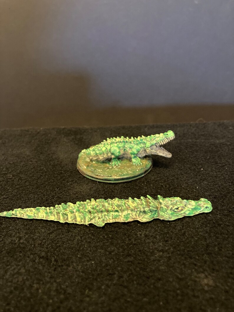 May include: Two miniature crocodile figurines. One is standing on a circular base, mouth open, and the other is lying flat. Both are painted in shades of green and brown, with detailed scales.