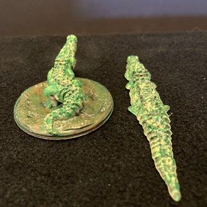 May include: Two green alligator figurines. One is atop a circular base, the other is a standalone figure. The alligators have textured skin and are painted in shades of green. The base has a weathered appearance.