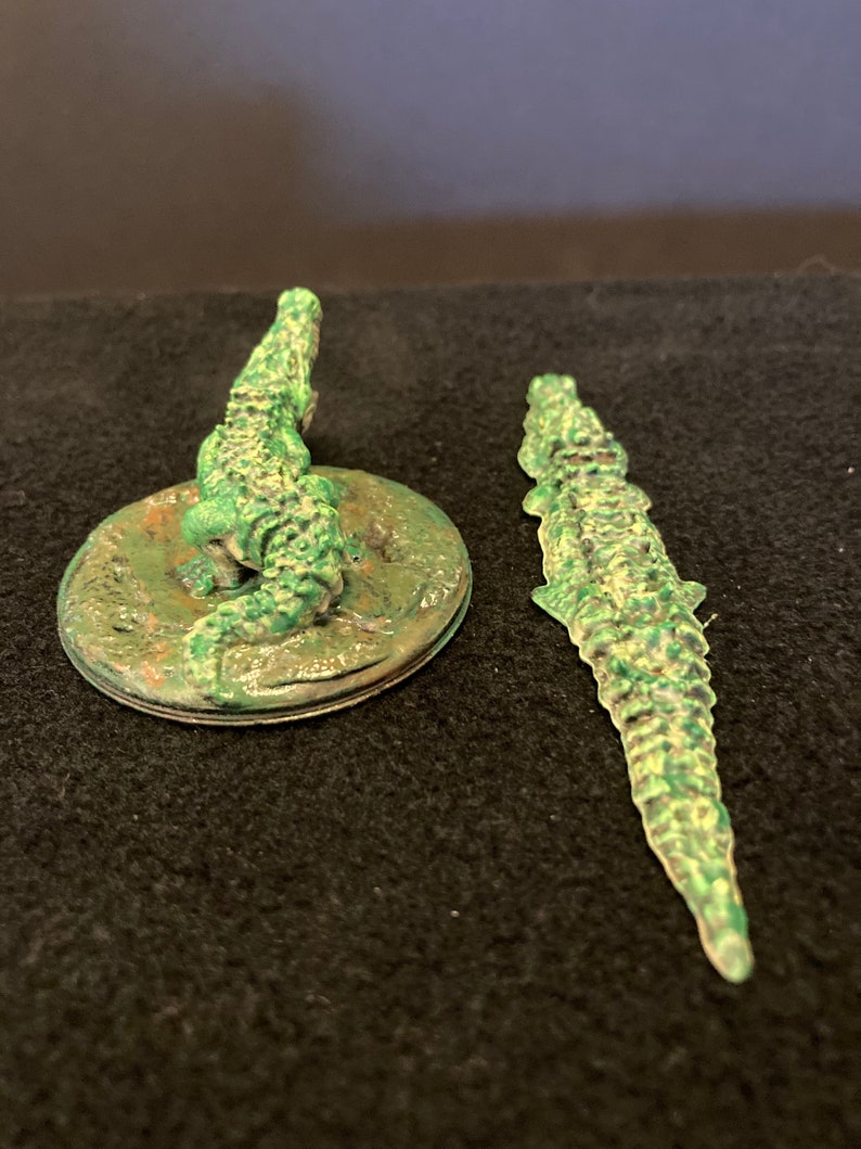 May include: Two green alligator figurines. One is atop a circular base, the other is a standalone figure. The alligators have textured skin and are painted in shades of green. The base has a weathered appearance.