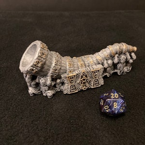 May include: A silver and gold detailed, 3D printed, dragon-shaped dice tower with a blue and gold 20-sided die in front of it.
