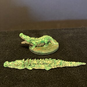 May include: Two miniature crocodile figurines. One is posed with its mouth open, the other is lying flat. Both are painted in shades of green and tan, with detailed scales and textures. The figurines are likely for tabletop gaming or display.