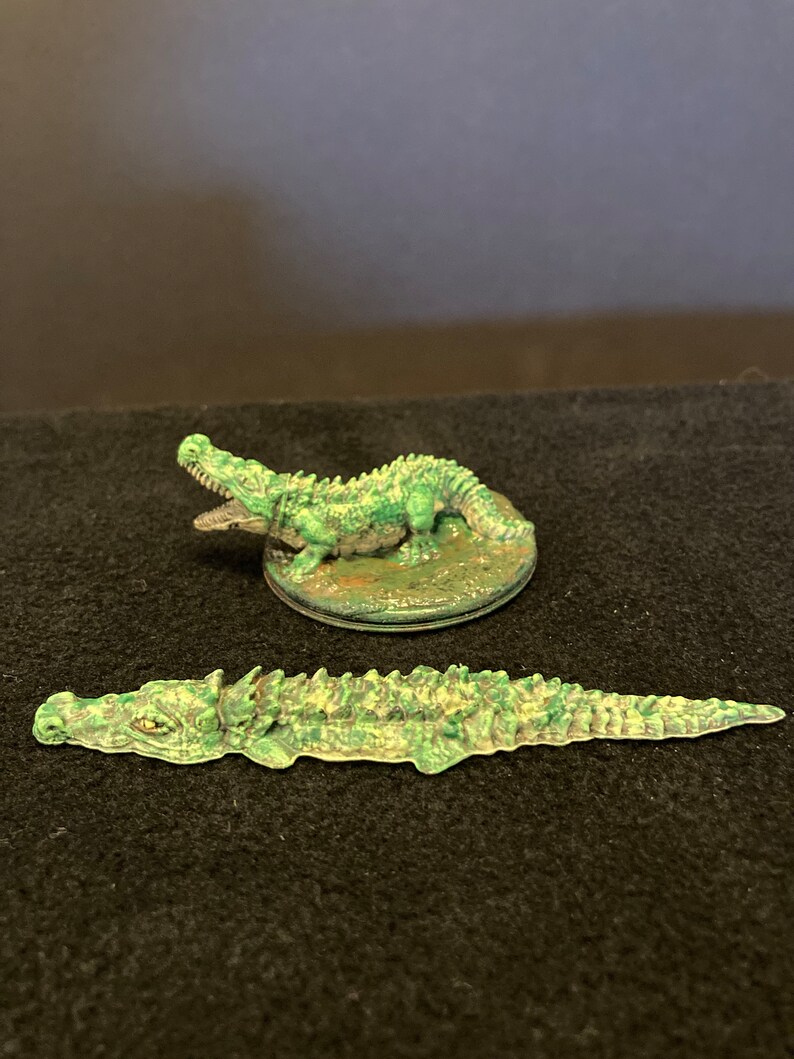 May include: Two miniature crocodile figurines. One is posed with its mouth open, the other is lying flat. Both are painted in shades of green and tan, with detailed scales and textures. The figurines are likely for tabletop gaming or display.