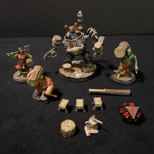 Op de afbeelding: Een set miniatuurfiguren voor een tabletop-spel, met een grote, metalen robot met een houthakkers-thema, twee kleinere figuren van orcs en een verscheidenheid aan houten boomstammen en gereedschap. De figuren zijn geschilderd in verschillende kleuren, waaronder groen, bruin en zilver.