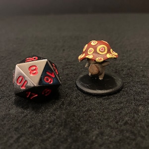 May include: A black metal twenty-sided die with red numbers and a miniature mushroom figurine with a red and yellow cap on a black base.