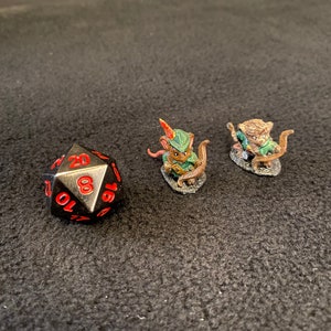 Puede incluir: Dos figuritas pintadas en miniatura de ratones vestidos de aventureros, uno con arco y flecha y el otro con una espada. Un dado negro de 20 caras con números rojos está en primer plano.