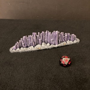 May include: A purple and gray crystal mountain terrain model for tabletop gaming. The model is made of plastic and has a textured surface. A black 20-sided die is in the foreground.