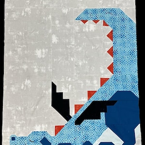 May include: A rectangular quilt featuring a pieced fabric design of a blue dragon with a textured scale pattern, black wings, and red triangular spikes along its back. The dragon is set against a light gray mottled background fabric.