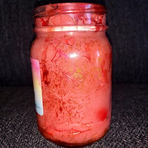 May include: A glass jar with a red, pink, and gold swirl design. The jar is filled with a red, pink, and gold substance. The jar has a white label with the text "The Good Witch" on it.
